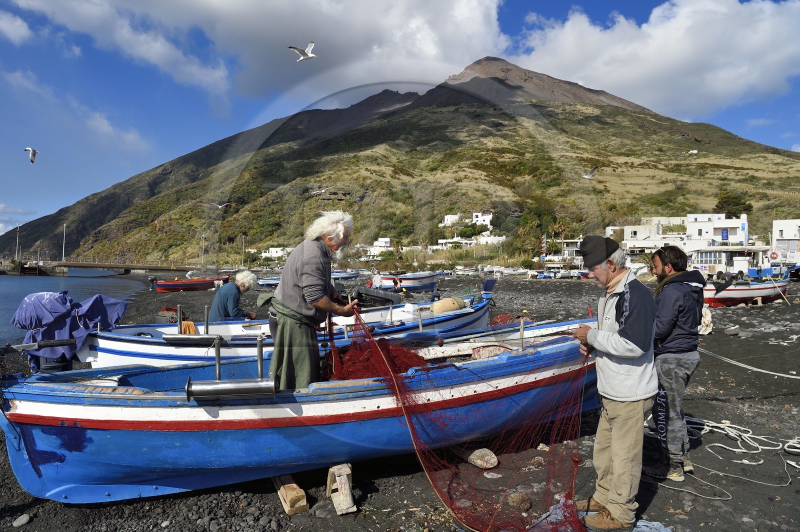 Italie, Sicile, iles Eoliennes, classées Patrimoine Mondial de l'UNESCO, ile de Stromboli, le pecheur Gaetano Cusolito réparant ses filets avec ses deux frères sur la plage de Scari et le volcan du Stromboli en arrière plan