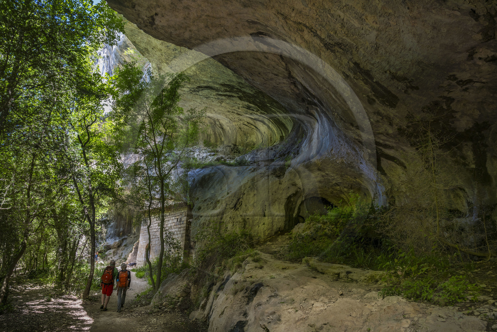 France, Vaucluse, Mont Ventoux Regional Natural Park, Monieux, Gorges de La Nesque, hikers walking under a rock ledge at the bottom of the canyon towards the 12th century Saint-Michel chapel