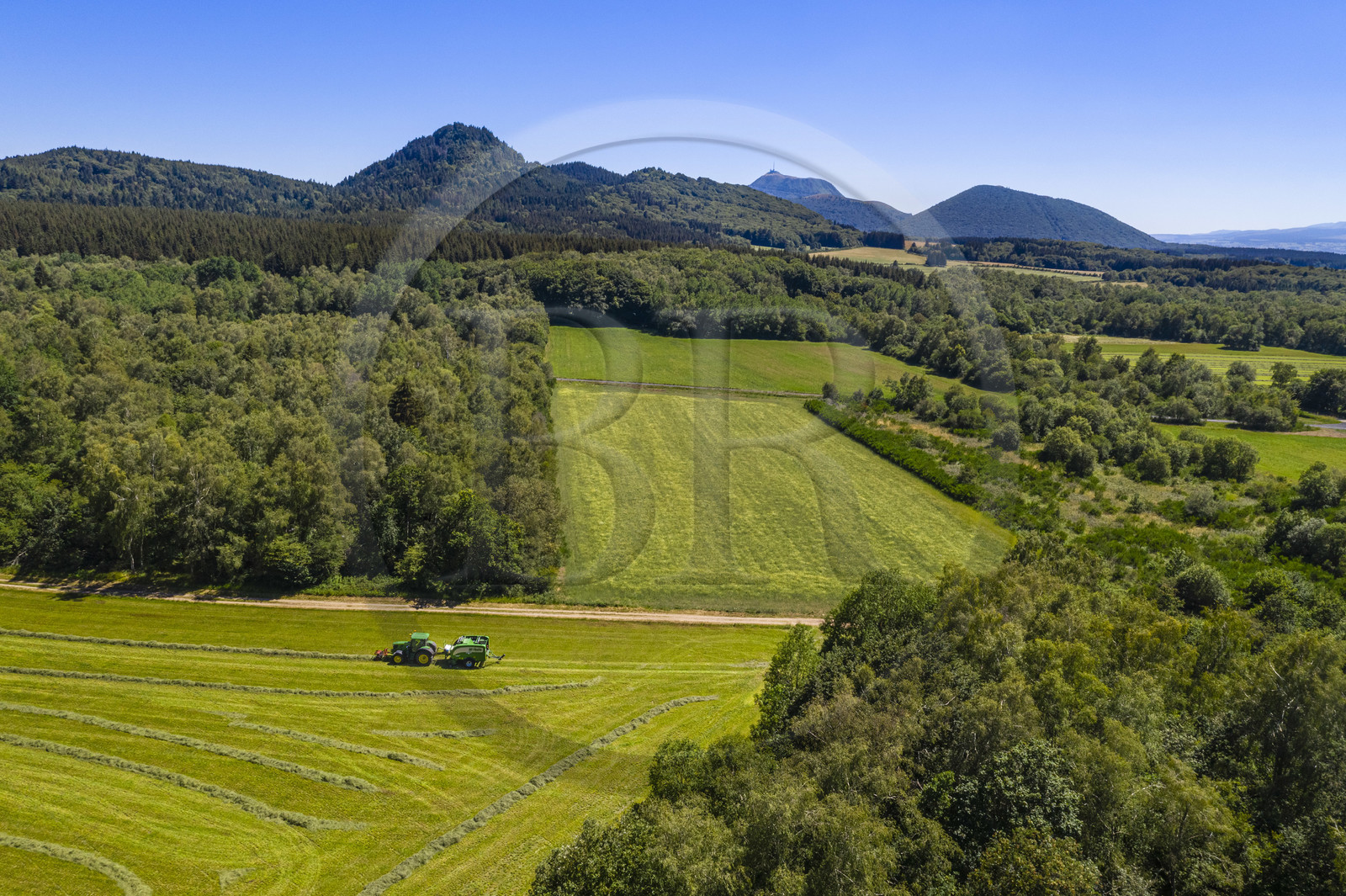 France, Puy-de-Dôme (63), Saint-Ours-les-Roches, hameau de Beauregard, tracteur récoltant le fourrage avec une presse enrubanneuse de balles de foin dans un champ, en arrière plan le volcan le Puy Chopine à gauche et le Puy de Dome à droite (vue aérienne)